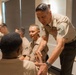 A senior U.S. Marine Corps non-commissioned officer, identifiable by his rank insignia and service ribbons, is leaning forward to shake hands with a seated Marine. The setting appears to be an indoor meeting or briefing room with multiple Marines seated at a table. The interaction suggests a formal or ceremonial greeting between a senior leader and junior enlisted personnel.