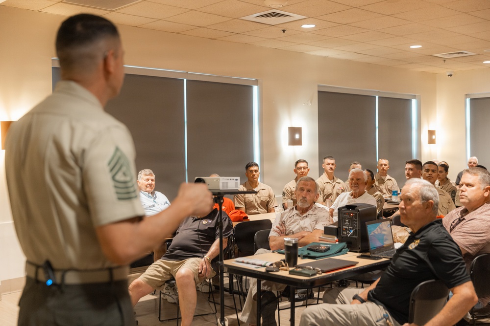A military briefing or presentation is taking place in a conference room, with a senior non-commissioned officer (identifiable by chevron rank insignia on sleeve) addressing a mixed audience of uniformed military personnel and civilians. The speaker stands at the front holding what appears to be a remote or pointer, facing the seated audience. The setting appears to be a modern military facility meeting room.