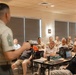 A military briefing or presentation is taking place in a conference room, with a senior non-commissioned officer (identifiable by chevron rank insignia on sleeve) addressing a mixed audience of uniformed military personnel and civilians. The speaker stands at the front holding what appears to be a remote or pointer, facing the seated audience. The setting appears to be a modern military facility meeting room.