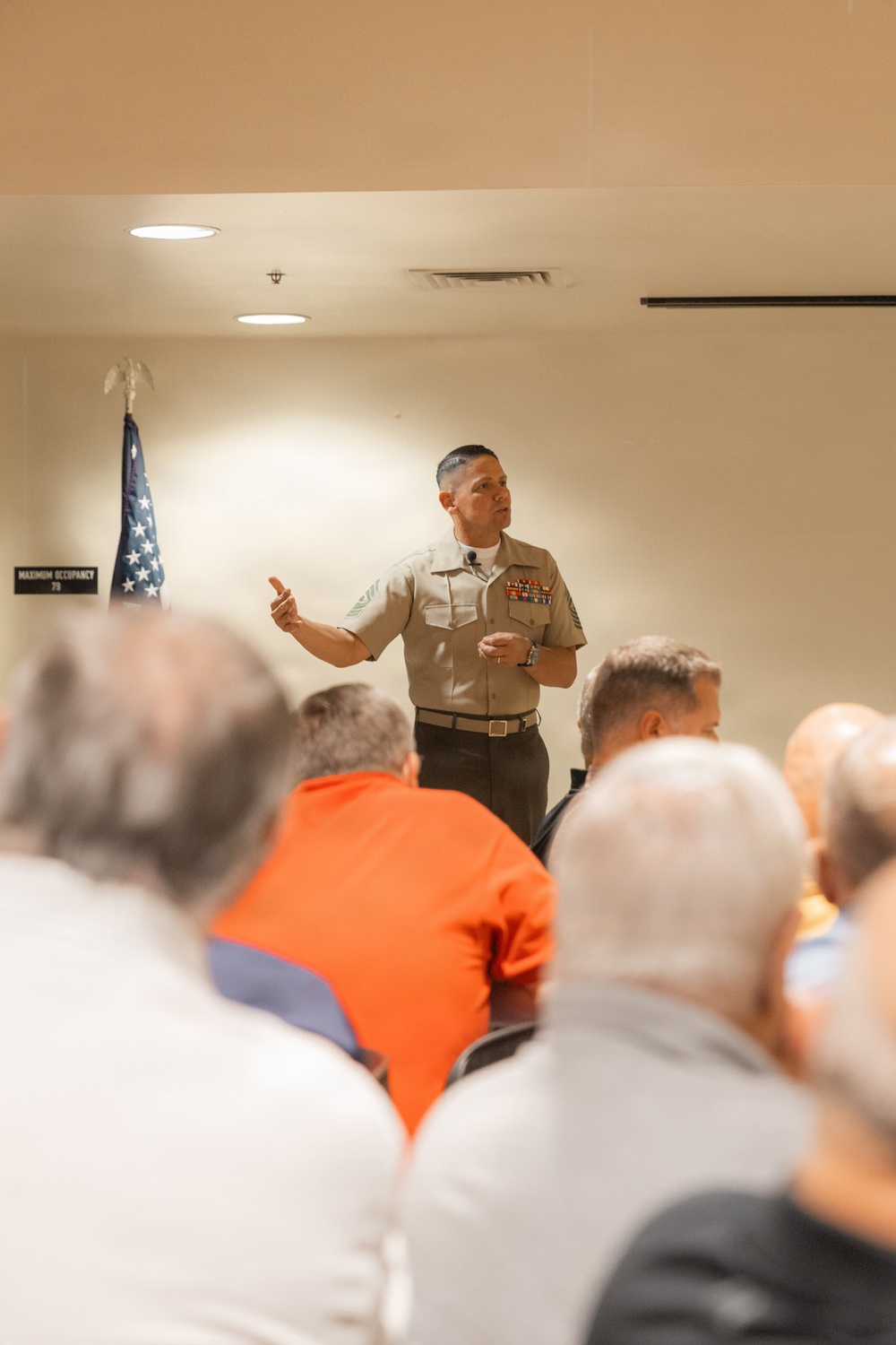 A U.S. Marine in service uniform (Charlie/Bravo uniform) is addressing a seated audience in an indoor meeting room. The speaker displays multiple ribbons/medals on his chest and appears to be a senior Non-Commissioned Officer based on his chevron insignia. An American flag is visible in the background alongside a maximum occupancy sign reading '70'.