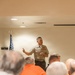 A U.S. Marine in service uniform (Charlie/Bravo uniform) is addressing a seated audience in an indoor meeting room. The speaker displays multiple ribbons/medals on his chest and appears to be a senior Non-Commissioned Officer based on his chevron insignia. An American flag is visible in the background alongside a maximum occupancy sign reading '70'.