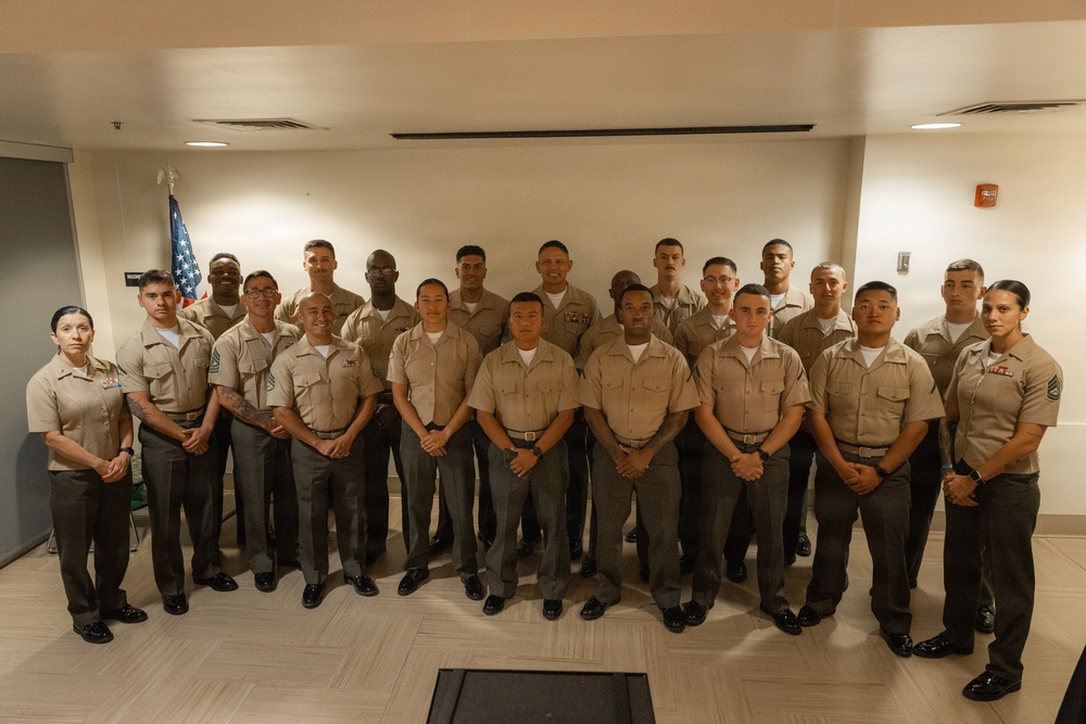 A formal group portrait of approximately 22-24 military personnel wearing U.S. Marine Corps service uniforms (tan/khaki shirts with olive/grey trousers). The group is arranged in three rows inside what appears to be a modern indoor facility. An American flag is partially visible in the background on the left side.