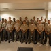 A formal group portrait of approximately 22-24 military personnel wearing U.S. Marine Corps service uniforms (tan/khaki shirts with olive/grey trousers). The group is arranged in three rows inside what appears to be a modern indoor facility. An American flag is partially visible in the background on the left side.