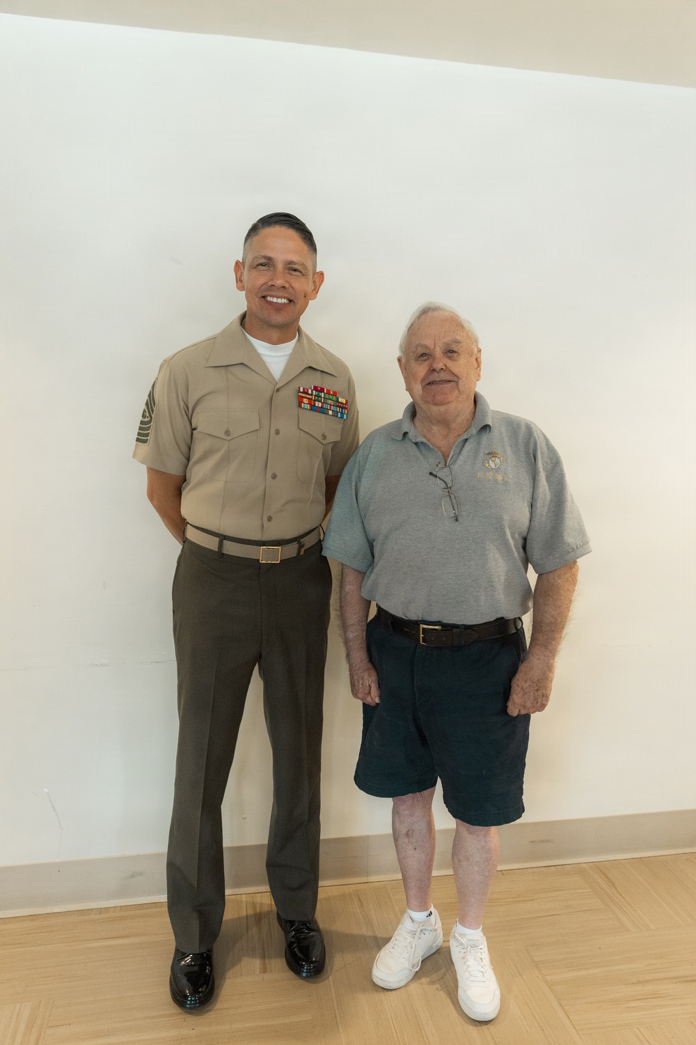Two men are posing together against a white wall on a light wood floor. The man on the left is wearing a U.S. Marine Corps Service 'Charlie' uniform with multiple service ribbons and a senior enlisted rank chevron on his sleeve. The older man on the right is wearing a gray polo shirt with what appears to be a military or veteran emblem, dark shorts, and white sneakers.