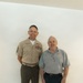 Two men are posing together against a white wall on a light wood floor. The man on the left is wearing a U.S. Marine Corps Service 'Charlie' uniform with multiple service ribbons and a senior enlisted rank chevron on his sleeve. The older man on the right is wearing a gray polo shirt with what appears to be a military or veteran emblem, dark shorts, and white sneakers.