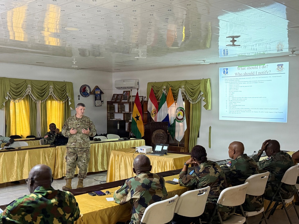 A U.S. military soldier in MultiCam/OCP camouflage uniform is delivering a briefing or training presentation to an audience of African military personnel in a conference room. The projected slide is titled 'What should I do? Who should I notify?' suggesting a training session on reporting procedures or conduct standards. The setting appears to be a military facility, likely in West Africa based on the visible national flags.