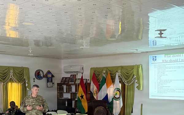 A U.S. military soldier in MultiCam/OCP camouflage uniform is delivering a briefing or training presentation to an audience of African military personnel in a conference room. The projected slide is titled 'What should I do? Who should I notify?' suggesting a training session on reporting procedures or conduct standards. The setting appears to be a military facility, likely in West Africa based on the visible national flags.