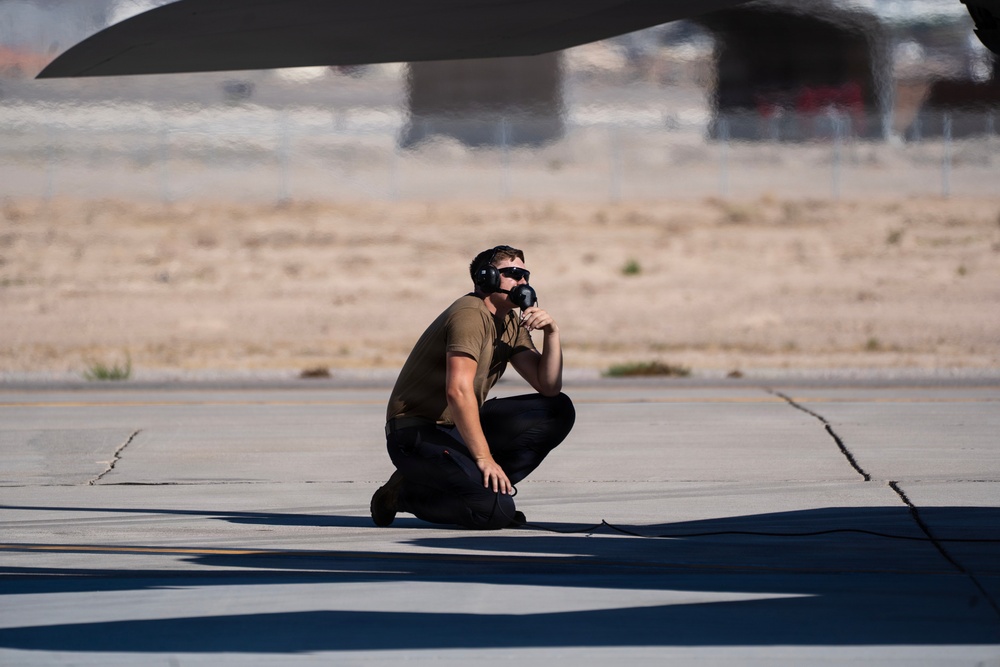 A military or aviation ground crew member is crouching on a tarmac beneath an aircraft wing, wearing hearing protection headset and sunglasses. He appears to be communicating via a headset microphone while monitoring or inspecting the aircraft. The setting is a desert airfield, likely a military air base based on the environment and attire.