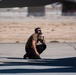 A military or aviation ground crew member is crouching on a tarmac beneath an aircraft wing, wearing hearing protection headset and sunglasses. He appears to be communicating via a headset microphone while monitoring or inspecting the aircraft. The setting is a desert airfield, likely a military air base based on the environment and attire.