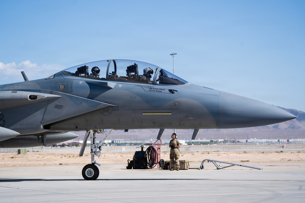 A military fighter jet, identifiable as an F-15 Eagle based on its distinctive twin-seat cockpit and airframe design, is positioned on a tarmac at what appears to be a desert air base. Two pilots in full flight gear are seated in the cockpit, while a ground crew member stands nearby next to support equipment. The setting suggests pre-flight or post-flight operations at a U.S. Air Force installation.