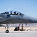 A military fighter jet, identifiable as an F-15 Eagle based on its distinctive twin-seat cockpit and airframe design, is positioned on a tarmac at what appears to be a desert air base. Two pilots in full flight gear are seated in the cockpit, while a ground crew member stands nearby next to support equipment. The setting suggests pre-flight or post-flight operations at a U.S. Air Force installation.