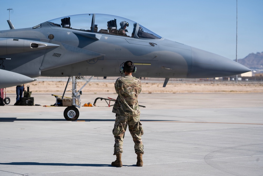 A military ground crew member in camouflage uniform and hearing protection stands on a flight line facing an F-15 Eagle fighter jet. The aircraft is positioned on the tarmac with a pilot visible in the cockpit, suggesting pre-flight or post-flight operations. The setting appears to be a desert air base, consistent with locations in the American Southwest.