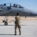 A military ground crew member in camouflage uniform and hearing protection stands on a flight line facing an F-15 Eagle fighter jet. The aircraft is positioned on the tarmac with a pilot visible in the cockpit, suggesting pre-flight or post-flight operations. The setting appears to be a desert air base, consistent with locations in the American Southwest.