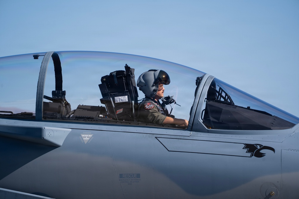 A military pilot is seated in the cockpit of a fighter jet, wearing a flight helmet and olive green flight suit with patches. The aircraft appears to be an F-15 Eagle, identifiable by the eagle emblem on the fuselage and the 'RESCUE' placard visible below the cockpit. The image is taken at close range on what appears to be a flight line or taxiway under clear skies.