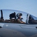 A military pilot is seated in the cockpit of a fighter jet, wearing a flight helmet and olive green flight suit with patches. The aircraft appears to be an F-15 Eagle, identifiable by the eagle emblem on the fuselage and the 'RESCUE' placard visible below the cockpit. The image is taken at close range on what appears to be a flight line or taxiway under clear skies.