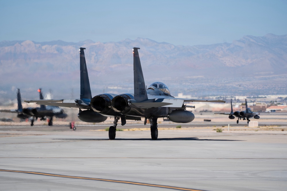 Multiple military jet aircraft are taxiing on a runway at what appears to be a desert air base. The foreground features a twin-tail fighter jet, identifiable as an F-15 Eagle, with at least two additional aircraft visible in the background. The setting includes arid terrain and mountain ranges consistent with a southwestern United States location.