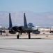 Multiple military jet aircraft are taxiing on a runway at what appears to be a desert air base. The foreground features a twin-tail fighter jet, identifiable as an F-15 Eagle, with at least two additional aircraft visible in the background. The setting includes arid terrain and mountain ranges consistent with a southwestern United States location.
