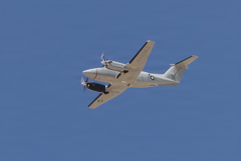 A U.S. Marines twin-turboprop aircraft is photographed in flight against a clear blue sky. The aircraft appears to be a UC-35 or similar Beechcraft King Air variant used by the United States Marine Corps. Both propellers are visibly spinning, indicating active flight.