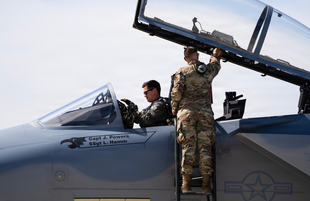 Two U.S. Air Force personnel are conducting pre-flight or post-flight procedures on a military jet aircraft. A pilot in a flight suit and sunglasses is seated in the cockpit, while a ground crew member in OCP (Operational Camouflage Pattern) uniform assists by operating the canopy. The aircraft fuselage displays the names 'Capt J. Powers' and 'SSgt L. Hamm' along with a unit insignia.