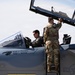 Two U.S. Air Force personnel are conducting pre-flight or post-flight procedures on a military jet aircraft. A pilot in a flight suit and sunglasses is seated in the cockpit, while a ground crew member in OCP (Operational Camouflage Pattern) uniform assists by operating the canopy. The aircraft fuselage displays the names 'Capt J. Powers' and 'SSgt L. Hamm' along with a unit insignia.