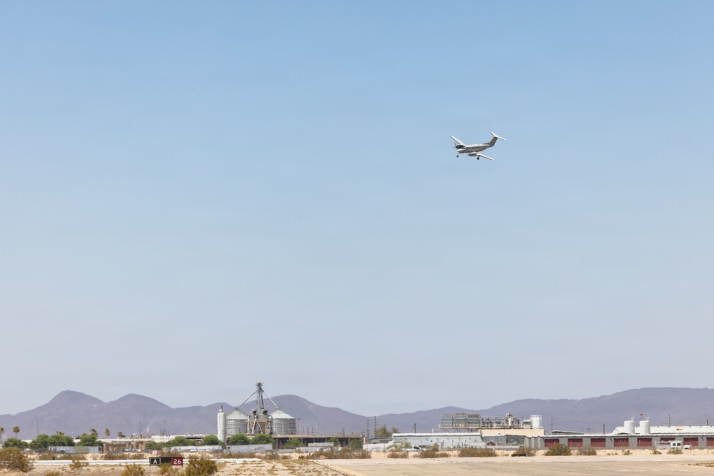 A small twin-engine turboprop aircraft is captured in flight over a desert airport environment. Industrial and airport infrastructure is visible along the horizon, backed by arid mountain ranges. The scene appears to be a small regional or general aviation airport in a desert region.