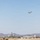 A small twin-engine turboprop aircraft is captured in flight over a desert airport environment. Industrial and airport infrastructure is visible along the horizon, backed by arid mountain ranges. The scene appears to be a small regional or general aviation airport in a desert region.