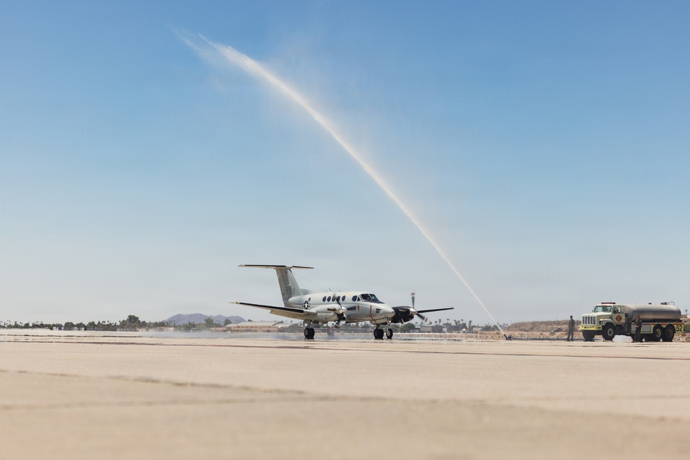 A military twin-turboprop aircraft, consistent with a Beechcraft King Air or C-12 variant, is receiving a water cannon salute on a tarmac. A yellow/olive military tanker truck (numbered 52) is positioned to the right, projecting a high arc of water over the aircraft. This ceremony typically marks a final flight or retirement of an aircraft or aircrew.