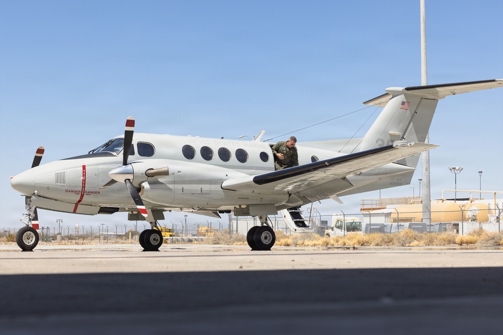 A white twin-engine turboprop aircraft, consistent with a Beechcraft King Air, is parked on a tarmac at what appears to be a military or government airfield. A person in olive/green flight suit is visible near the aircraft's door or wing area, likely conducting a pre-flight inspection or boarding. The aircraft displays 'DANGER' markings in red and features a US flag on the tail.