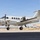 A white twin-engine turboprop aircraft, consistent with a Beechcraft King Air, is parked on a tarmac at what appears to be a military or government airfield. A person in olive/green flight suit is visible near the aircraft's door or wing area, likely conducting a pre-flight inspection or boarding. The aircraft displays 'DANGER' markings in red and features a US flag on the tail.