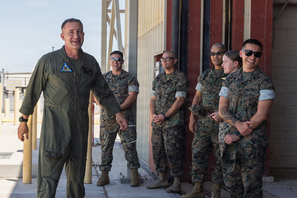 A military officer in an olive green flight suit stands to the left, smiling and apparently speaking to a group of five Marines in MARPAT camouflage uniforms. The group appears relaxed and engaged in a lighthearted interaction, with several individuals laughing or smiling. The setting appears to be an outdoor area adjacent to a military hangar or maintenance facility.
