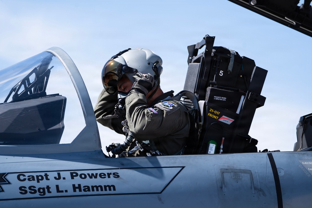 A military pilot in full flight gear, including a helmet with visor and oxygen mask, is seated in the cockpit of a fighter jet. The aircraft fuselage displays the names 'Capt J. Powers' and 'SSgt L. Hamm' in black lettering. The pilot appears to be conducting pre-flight checks or adjusting equipment before a mission.
