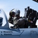 A military pilot in full flight gear, including a helmet with visor and oxygen mask, is seated in the cockpit of a fighter jet. The aircraft fuselage displays the names 'Capt J. Powers' and 'SSgt L. Hamm' in black lettering. The pilot appears to be conducting pre-flight checks or adjusting equipment before a mission.