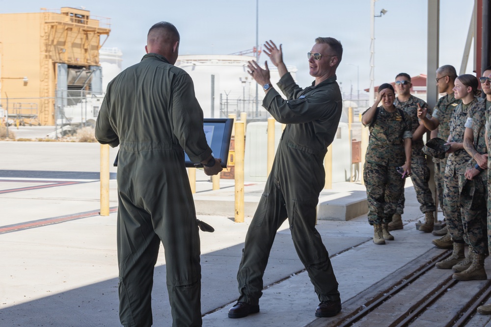 Two individuals in olive green flight suits are engaged in what appears to be a military ceremony or presentation, with one person holding a framed item and the other gesturing expressively. A group of military personnel in camouflage uniforms stands watching along the right side. The setting appears to be an outdoor area of a military airbase or installation.