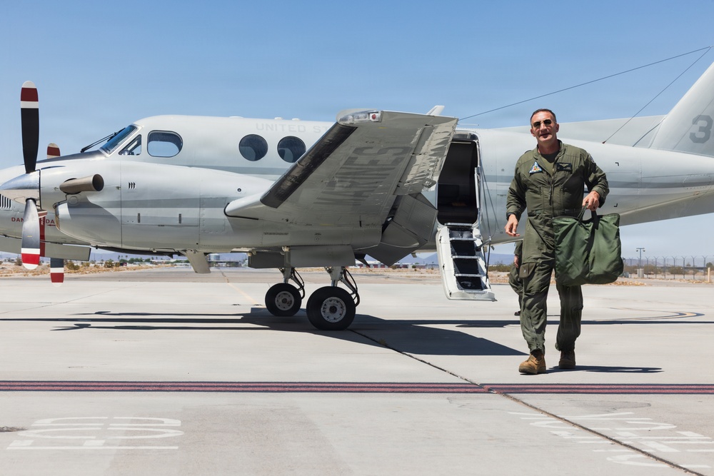 A military aviator in an olive green flight suit walks away from a light gray twin-engine turboprop aircraft on a military airfield tarmac. The aircraft door is open with steps deployed, suggesting the pilot has just disembarked. The setting appears to be a desert military installation based on the arid terrain visible in the background.