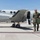 A military aviator in an olive green flight suit walks away from a light gray twin-engine turboprop aircraft on a military airfield tarmac. The aircraft door is open with steps deployed, suggesting the pilot has just disembarked. The setting appears to be a desert military installation based on the arid terrain visible in the background.
