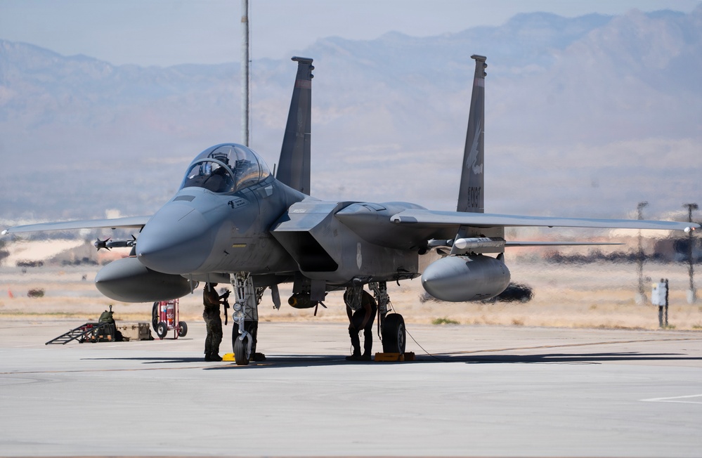 A U.S. Air Force F-15 Eagle fighter jet is parked on a flight line apron, likely at a desert air base. Ground crew personnel are performing pre-flight or post-flight maintenance checks beneath the aircraft. The background reveals arid terrain and mountain ranges consistent with a southwestern U.S. installation.