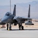 A U.S. Air Force F-15 Eagle fighter jet is parked on a flight line apron, likely at a desert air base. Ground crew personnel are performing pre-flight or post-flight maintenance checks beneath the aircraft. The background reveals arid terrain and mountain ranges consistent with a southwestern U.S. installation.