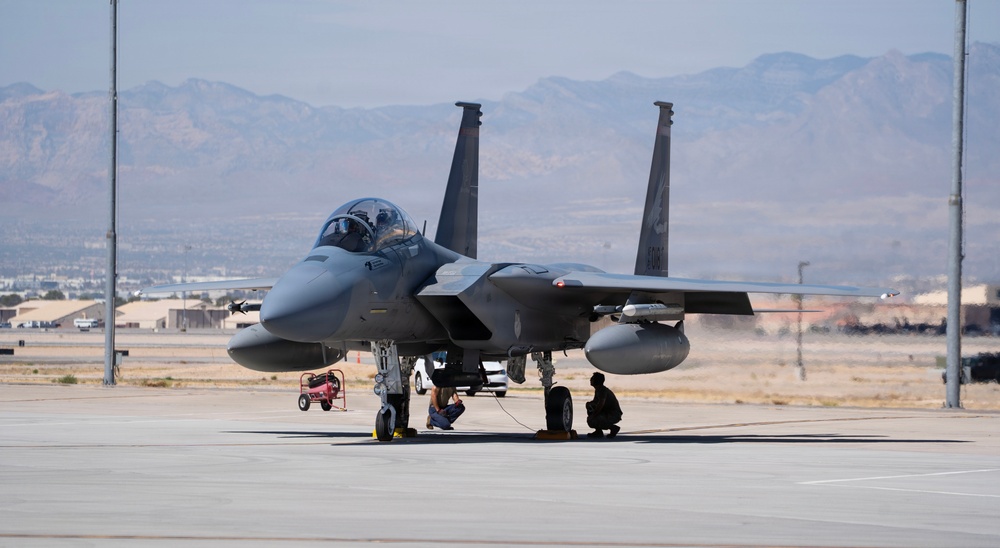 A twin-engine fighter jet, identifiable as an F-15 Eagle, is parked on a military airfield tarmac with ground crew personnel crouching beneath it performing pre-flight or post-flight checks. The aircraft is armed with visible missiles and external fuel tanks. The setting appears to be a desert military installation, with arid mountains visible in the background.