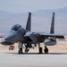 A twin-engine fighter jet, identifiable as an F-15 Eagle, is parked on a military airfield tarmac with ground crew personnel crouching beneath it performing pre-flight or post-flight checks. The aircraft is armed with visible missiles and external fuel tanks. The setting appears to be a desert military installation, with arid mountains visible in the background.