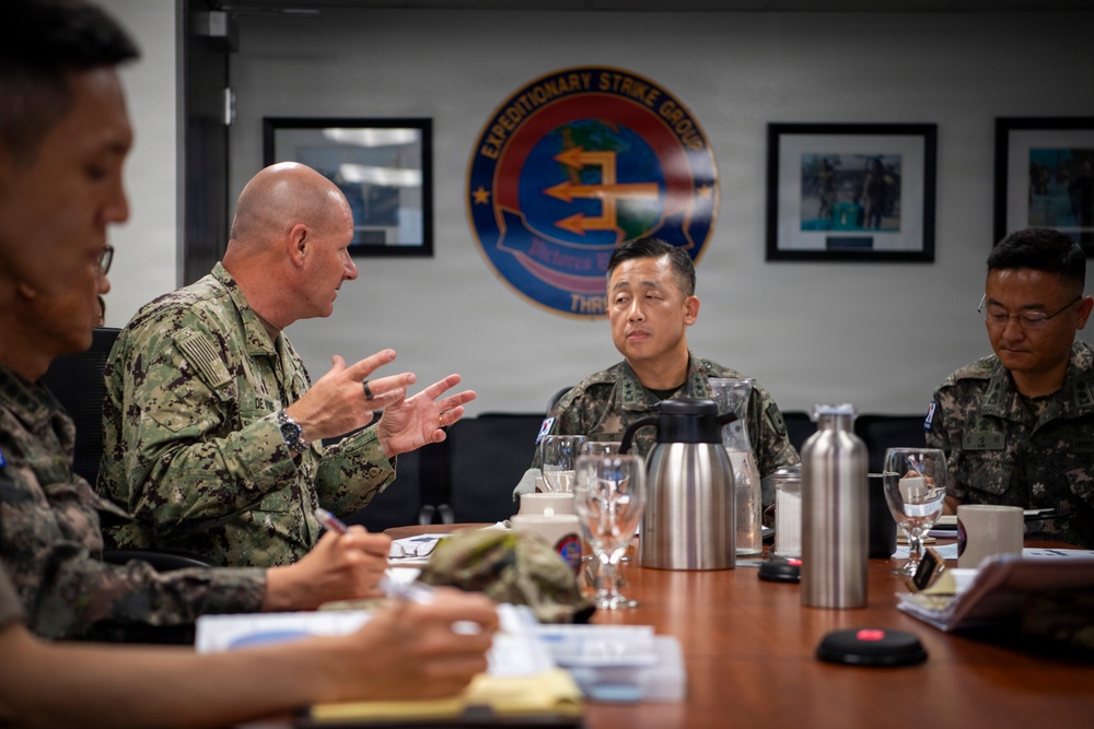 A military meeting is taking place in a conference room bearing the insignia of Expeditionary Strike Group 3 (ESG-3). A bald U.S. Navy officer in digital camouflage uniform is speaking and gesturing with his hands to a group of what appear to be Republic of Korea (ROK) military personnel. The setting suggests a bilateral military coordination or liaison meeting.