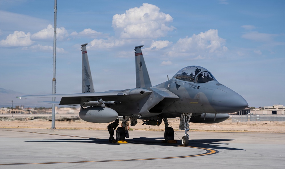 A U.S. Air Force F-15 Eagle fighter jet sits on a tarmac at what appears to be a desert air base, with tail number '013' visible on the twin vertical stabilizers. A ground crew member is performing a pre-flight or post-flight inspection near the main landing gear. The aircraft is armed with external munitions and appears to be in active operational status.