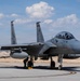 A U.S. Air Force F-15 Eagle fighter jet sits on a tarmac at what appears to be a desert air base, with tail number '013' visible on the twin vertical stabilizers. A ground crew member is performing a pre-flight or post-flight inspection near the main landing gear. The aircraft is armed with external munitions and appears to be in active operational status.