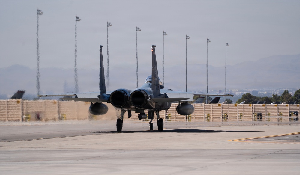 A military fighter jet, identifiable as an F-15 Eagle, is taxiing on a flight line at what appears to be a U.S. Air Force base, likely Nellis AFB based on the desert terrain and urban skyline visible in the background. The aircraft is viewed head-on, showing its twin-engine configuration and twin vertical stabilizers. The tarmac surface shows heat shimmer or moisture reflection, and concrete revetments (blast walls) are visible along the flight line.