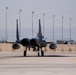 A military fighter jet, identifiable as an F-15 Eagle, is taxiing on a flight line at what appears to be a U.S. Air Force base, likely Nellis AFB based on the desert terrain and urban skyline visible in the background. The aircraft is viewed head-on, showing its twin-engine configuration and twin vertical stabilizers. The tarmac surface shows heat shimmer or moisture reflection, and concrete revetments (blast walls) are visible along the flight line.