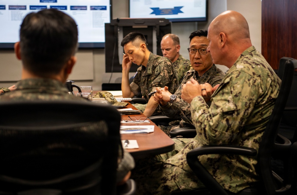 A military briefing or meeting is taking place around a conference table, involving personnel in at least two distinct camouflage uniforms — one consistent with US Navy Working Uniform (NWU) and others consistent with Republic of Korea (ROK) Army uniforms. The participants appear engaged in active discussion, with presentation screens visible in the background displaying briefing slides. The setting suggests a joint military coordination or planning session.
