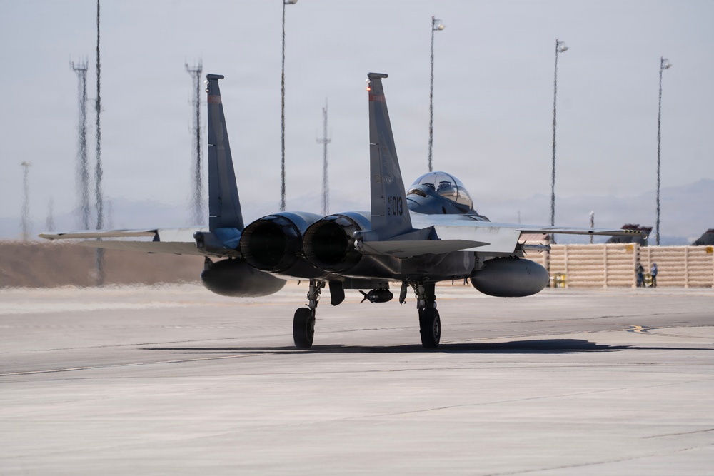 A twin-engine military fighter jet, identifiable as an F-15 Eagle, is taxiing on a concrete flight line at what appears to be a desert air base. The aircraft bears the tail number '013' and is viewed from a front-facing angle, showing its twin vertical stabilizers, dual engine exhausts, and external fuel tanks. The surrounding environment includes lighting poles, blast walls (HESCOs), and ground personnel visible in the background.