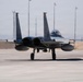 A twin-engine military fighter jet, identifiable as an F-15 Eagle, is taxiing on a concrete flight line at what appears to be a desert air base. The aircraft bears the tail number '013' and is viewed from a front-facing angle, showing its twin vertical stabilizers, dual engine exhausts, and external fuel tanks. The surrounding environment includes lighting poles, blast walls (HESCOs), and ground personnel visible in the background.