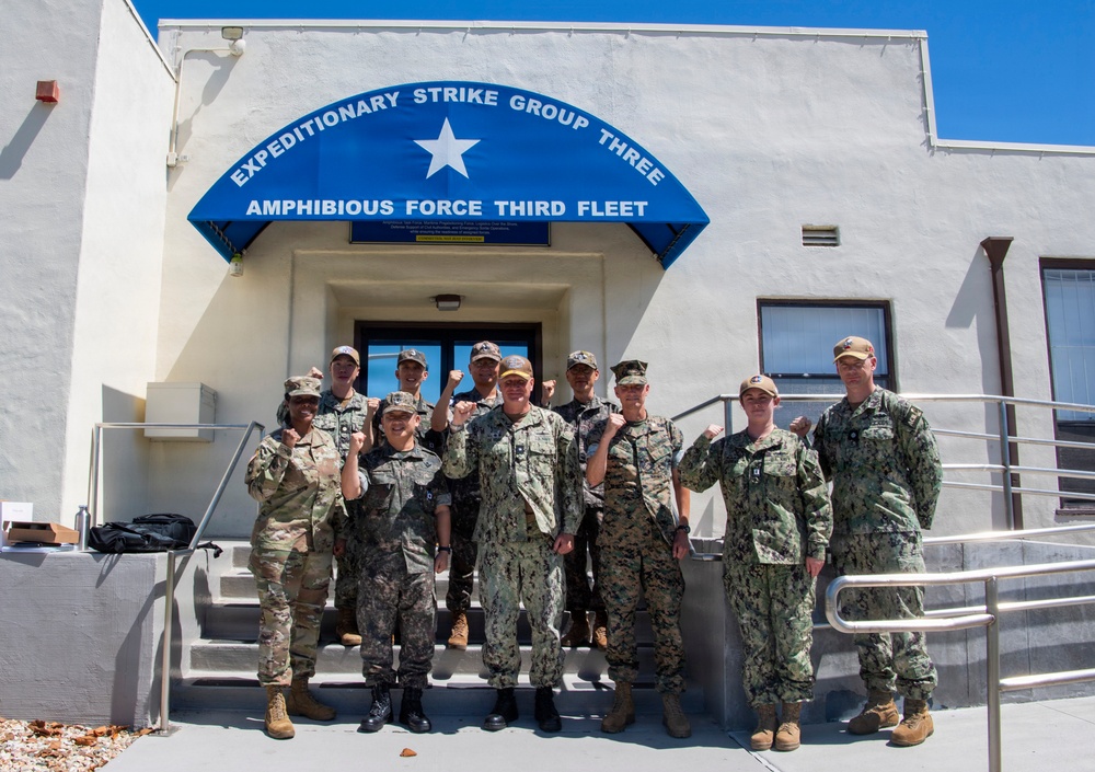 A group of approximately 12 military personnel pose for a group photo on the steps of the Expeditionary Strike Group Three / Amphibious Force Third Fleet headquarters building. Several members are raising their fists in a celebratory or solidarity gesture. The group includes members wearing multiple uniform types, indicating a joint or multi-service gathering.