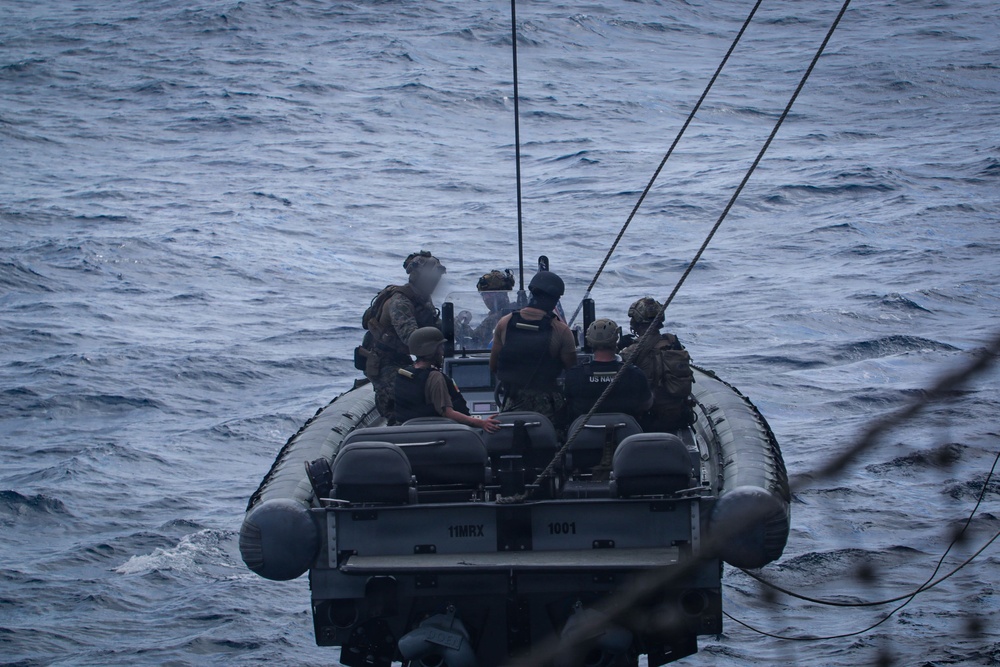 A military rigid inflatable boat (RIB) designated '11MRX 1001' is underway on open ocean waters, carrying multiple armed personnel in tactical gear. The personnel appear to be U.S. Navy and/or Marine Corps operators based on visible markings and equipment. The boat is equipped with communications antennas and appears to be connected to a larger vessel via lines visible in the foreground.