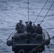 A military rigid inflatable boat (RIB) designated '11MRX 1001' is underway on open ocean waters, carrying multiple armed personnel in tactical gear. The personnel appear to be U.S. Navy and/or Marine Corps operators based on visible markings and equipment. The boat is equipped with communications antennas and appears to be connected to a larger vessel via lines visible in the foreground.